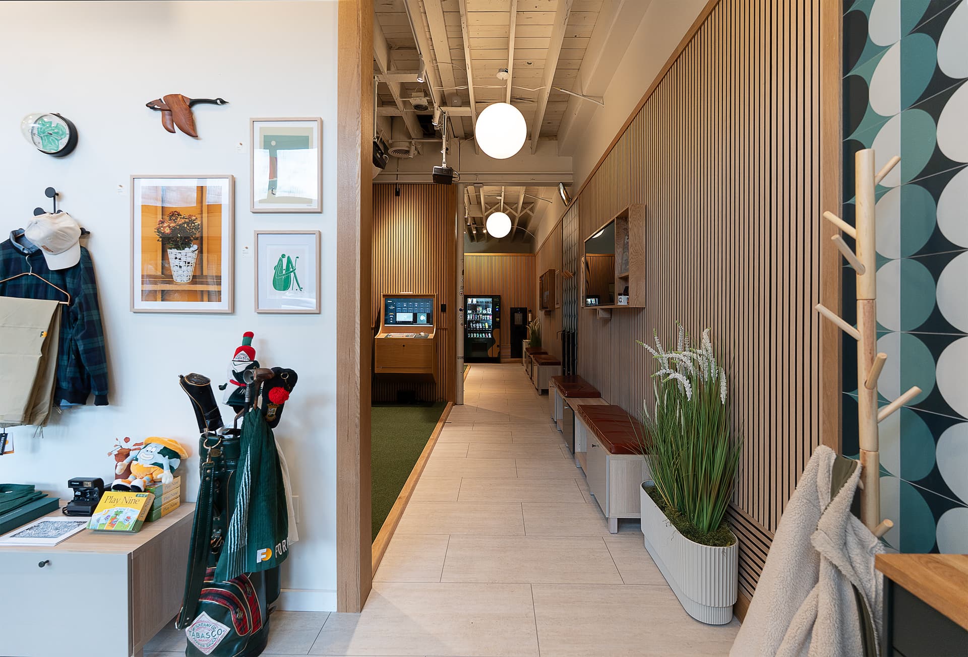 Hour Golf interior hallway with hanging jackets, golf bags, framed artwork, and a wood-slat wall leading back to the simulator bays
