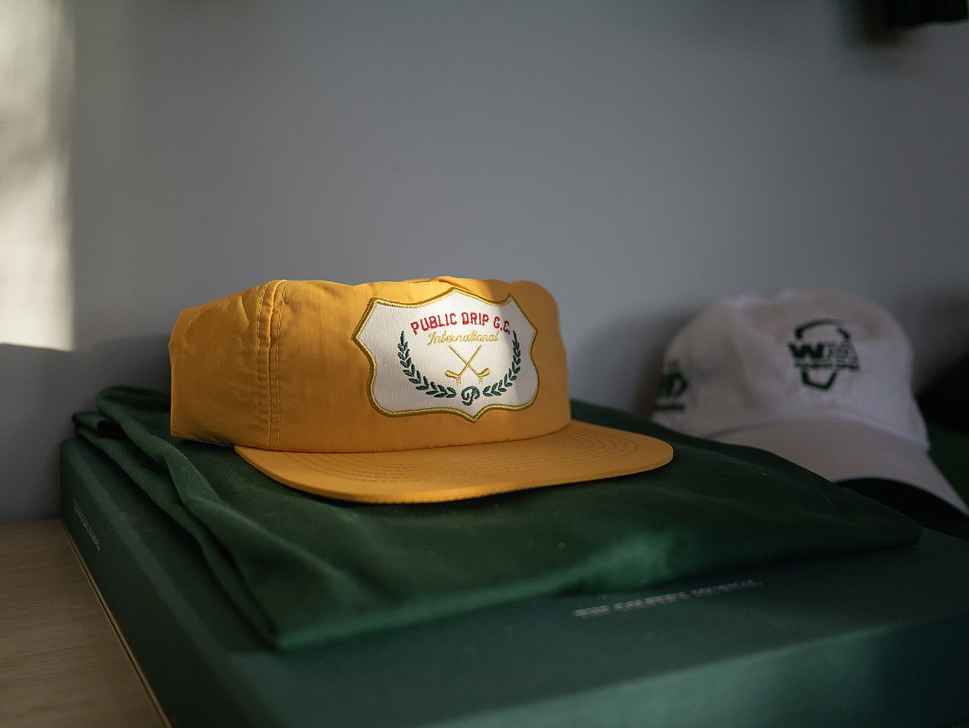 A yellow Public Drip GC golf cap and a white Hour Golf cap resting on a folded green jacket